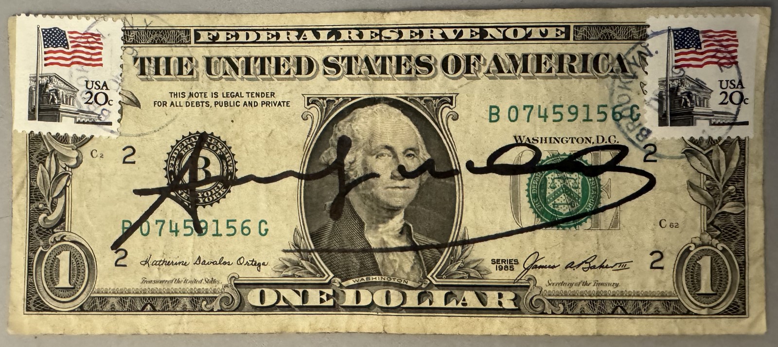 Andy WARHOL (1928-1987), Attribué à. Billet de 1 dollar Américain. Signé « Andy Warhol » au marqueur. Billet datant de 1985 et portant deux timbres de 20ct portant le cachet US Air Mail. Cachet de la signature au dos. Dimensions : 6,6 x 15,7 cm.