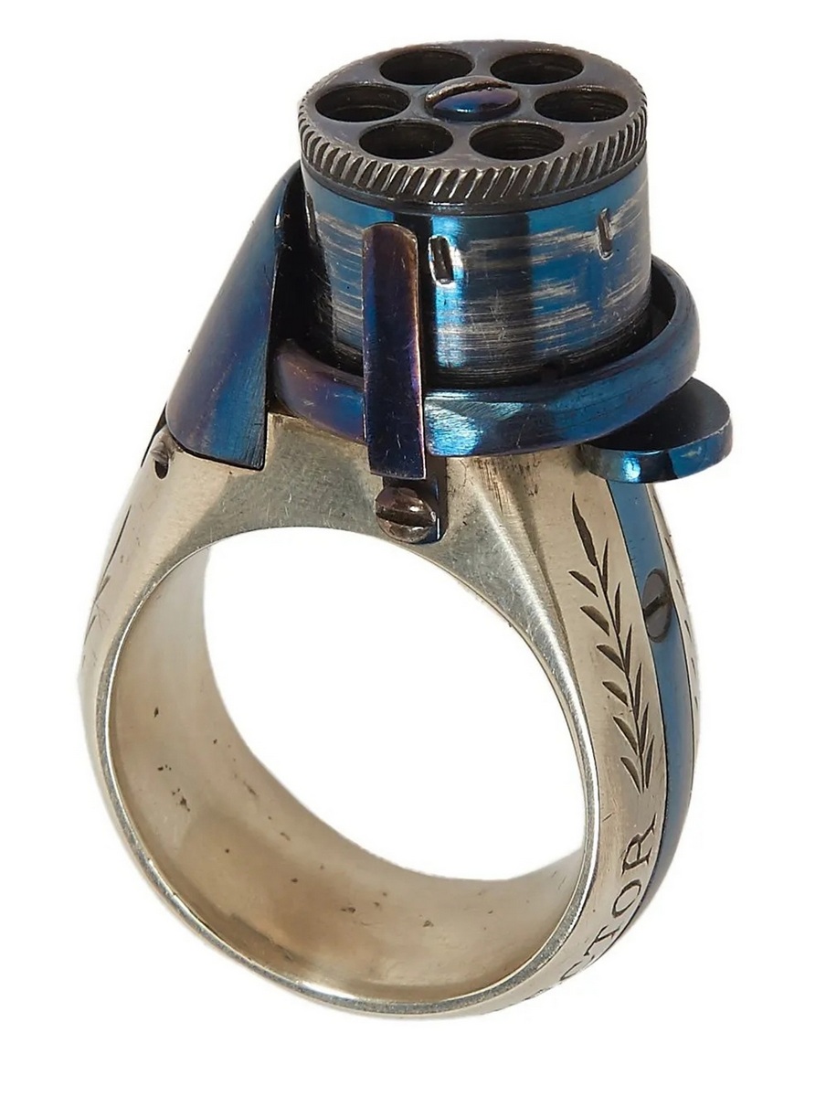 BAGUE RÉVOLVER, « le petit Protector », à broche, 6 coups, calibre 4 mm. Barillet bleu, guilloché sur le bord pour être tourné à la main. Détente latérale. Anneau en maillechort gravé de feuillages et marqué « LE PETIT PROTECTOR » XXe siècle. Dans son coffret en cuir décoré au petit fer, avec plaque en argent gravée « Le petit protector », garni à l’intérieur de velours. Avec sa boîte d’origine (6,7 x 8,2 , 2 cm) , 6 balles de plomb et un tournevis. Complet.