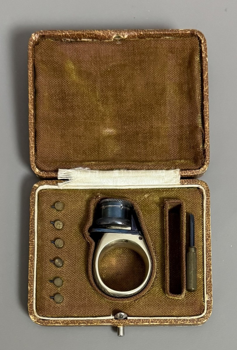 BAGUE RÉVOLVER, « le petit Protector », à broche, 6 coups, calibre 4 mm. Barillet bleu, guilloché sur le bord pour être tourné à la main. Détente latérale. Anneau en maillechort gravé de feuillages et marqué « LE PETIT PROTECTOR » XXe siècle. Dans son coffret en cuir décoré au petit fer, avec plaque en argent gravée « Le petit protector », garni à l’intérieur de velours. Avec sa boîte d’origine (6,7 x 8,2 , 2 cm) , 6 balles de plomb et un tournevis. Complet. - Image 2