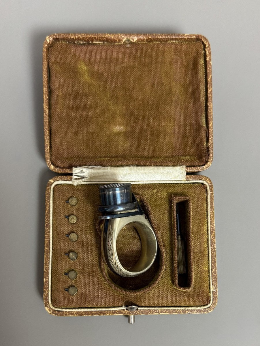 BAGUE RÉVOLVER, « le petit Protector », à broche, 6 coups, calibre 4 mm. Barillet bleu, guilloché sur le bord pour être tourné à la main. Détente latérale. Anneau en maillechort gravé de feuillages et marqué « LE PETIT PROTECTOR » XXe siècle. Dans son coffret en cuir décoré au petit fer, avec plaque en argent gravée « Le petit protector », garni à l’intérieur de velours. Avec sa boîte d’origine (6,7 x 8,2 , 2 cm) , 6 balles de plomb et un tournevis. Complet. - Image 6
