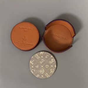 LANCEL & SALVADOR DALI (1904–1989). « Le Daligramme ». Circa 1980.  Ensemble de six sous-verres en cuir de vachette et tissu embossé, édités par la maison Lancel, marqués « Le Daligramme ». Dans son emboitage d’origine. Embossé sous l’emboitage « French légèreté depuis 1875 ». Diamètre : 10 cm.